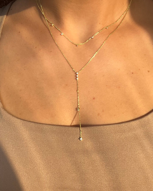 A close-up of a person's neck and upper chest wearing a delicate gold Y-necklace with a long drop chain and small clear stones, paired with a matching short chain necklace.