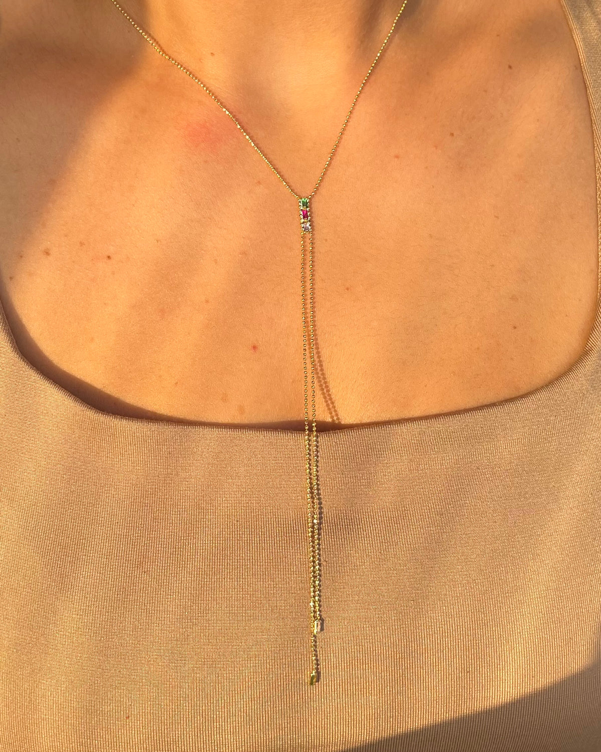 A close-up of a model wearing a delicate gold Y-necklace with a short drop chain and a small colorful gemstone accent at the center, paired with a beige scoop-neck top.