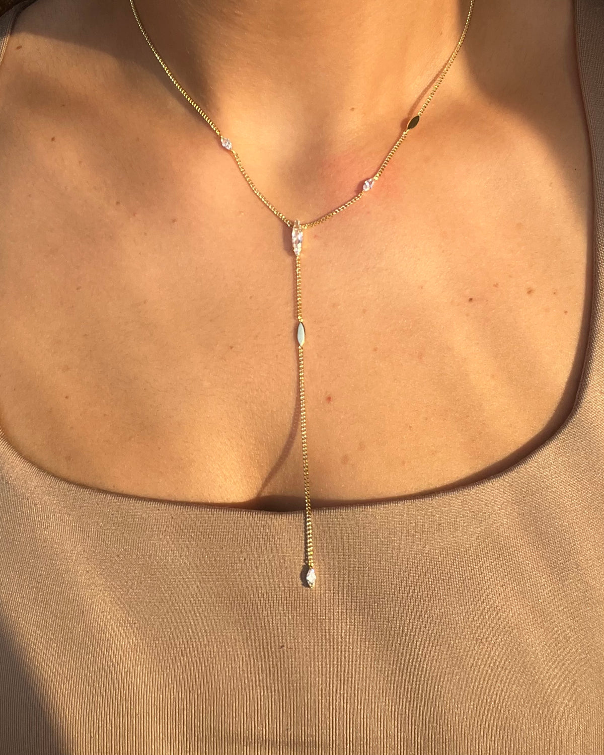 A delicate gold Y-necklace with small clear stones and a central drop ending in a teardrop-shaped clear stone, worn on a model's neck over a beige top.