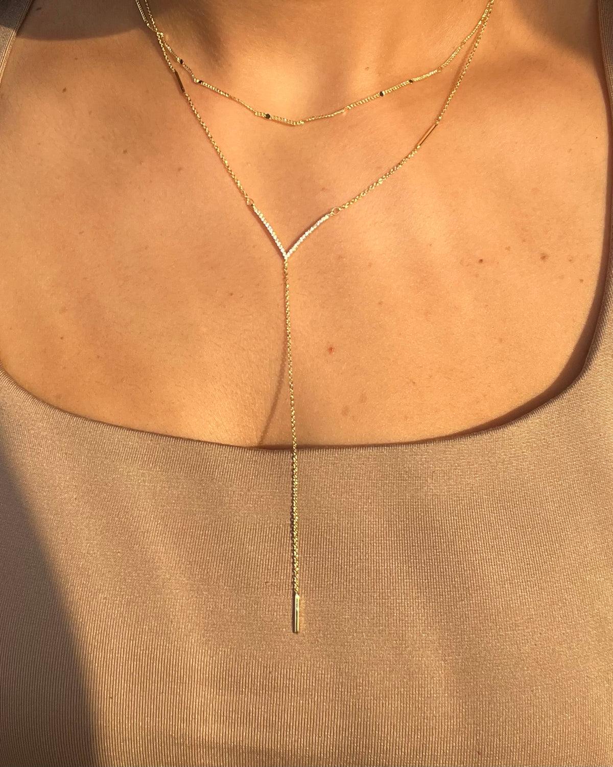 A close-up of a model's chest wearing a delicate gold Y-shaped lariat necklace over a beige scoop-neck top.