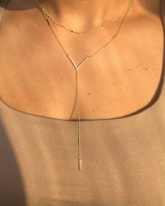 A close-up of a model's chest wearing a delicate gold Y-shaped lariat necklace over a beige scoop-neck top.