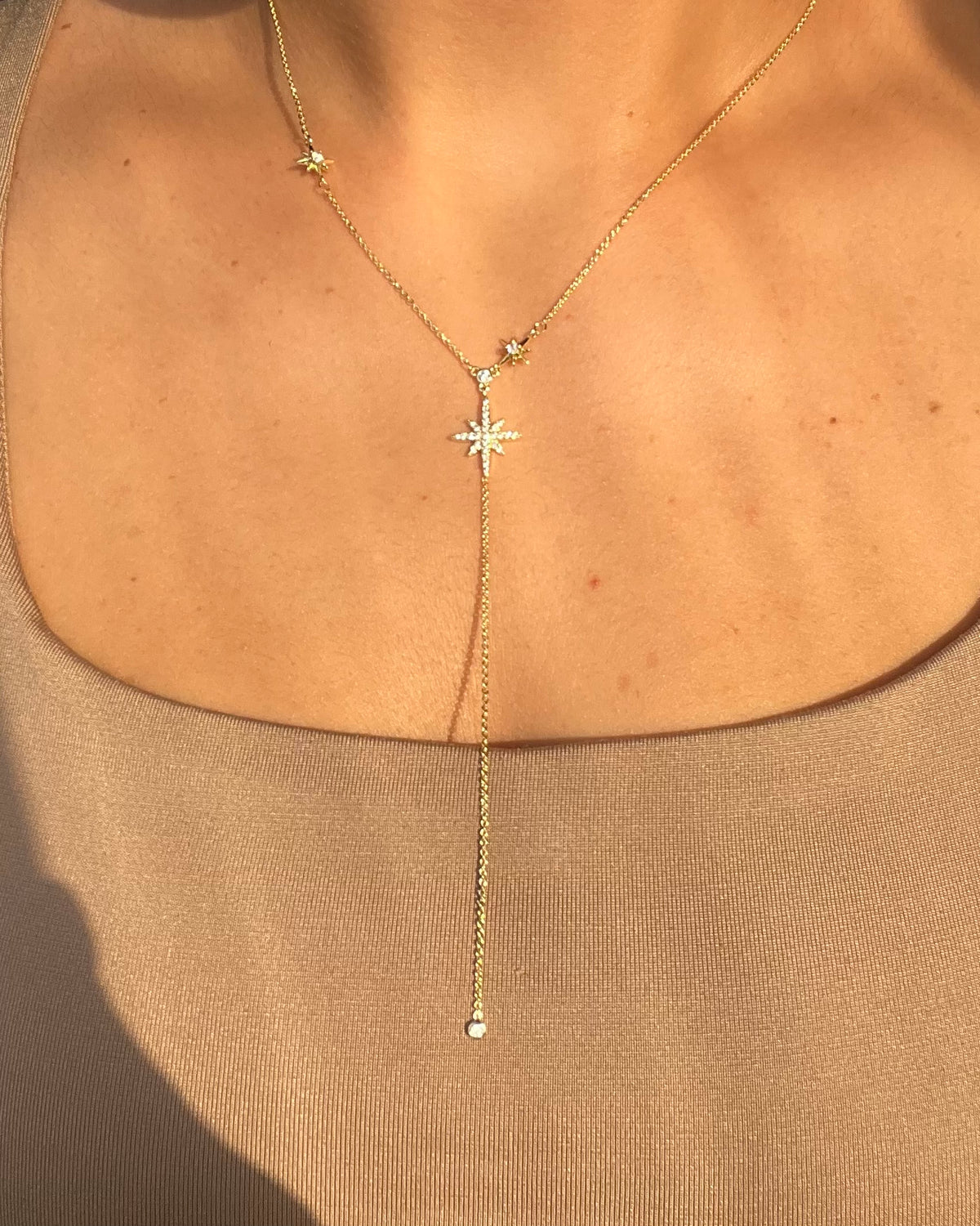 A gold-toned lariat necklace with a starburst pendant and small star accents, shown worn on a model's neck over a beige top.